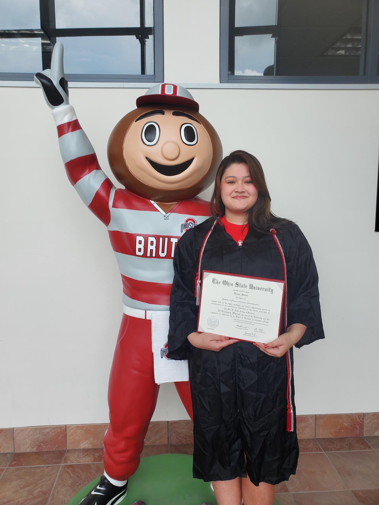 Female smiling displaying a diploma while standing next to Brutus Buckeye.