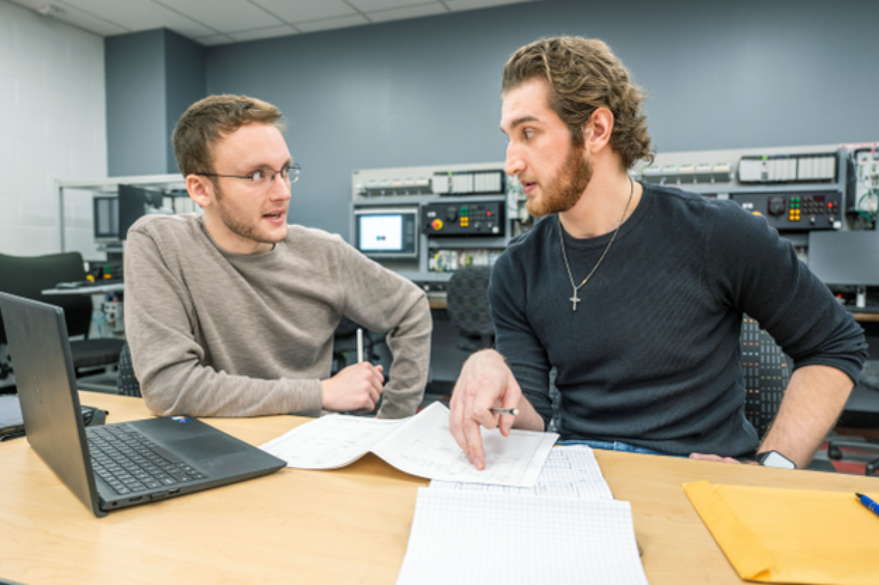 two male students sitting at a laptop speaking with each other