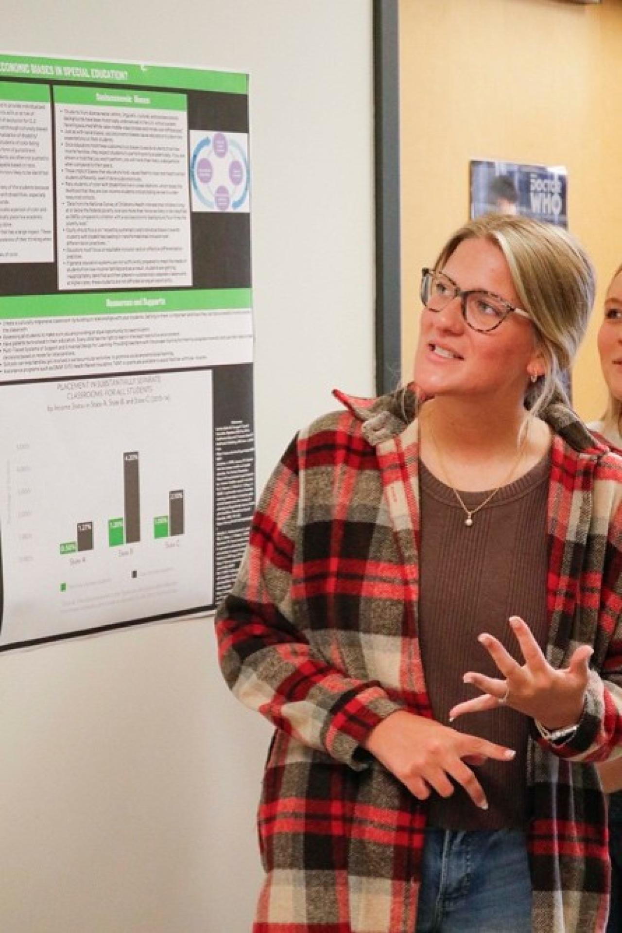 Photograph of a woman presenting a research poster featuring a bar chart with green and black bars, text sections, and a circular diagram. The poster appears to discuss a study or survey, with the bar chart highlighting key data points and comparisons, while the presenter gestures to emphasize her explanation.