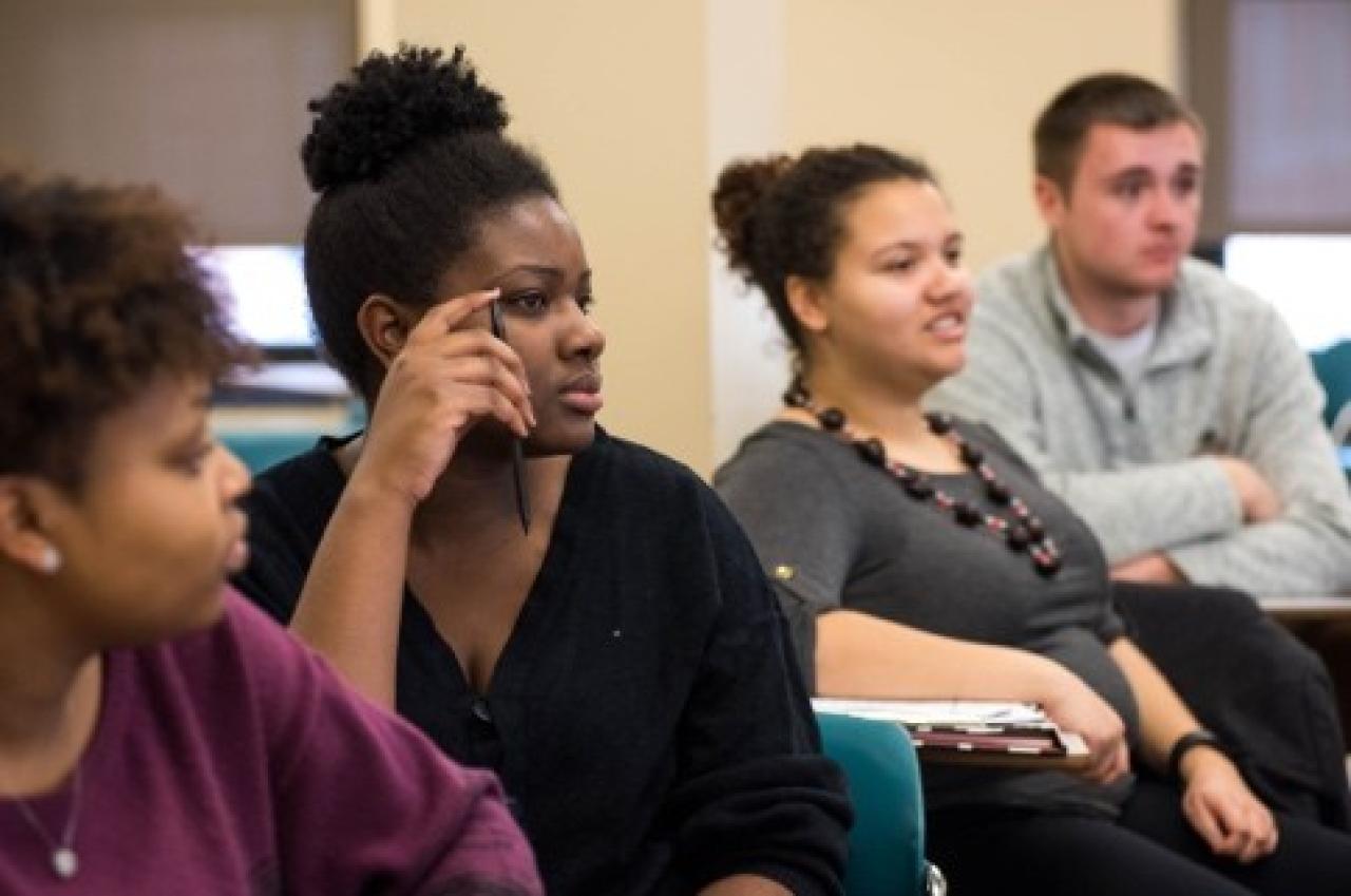 Photograph of four students seated in a classroom setting, attentively listening and engaged in a discussion or lecture. The diverse group includes two women and one man, with one woman holding a pen near her face, indicating active participation.