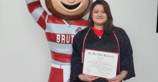 Female smiling displaying a diploma while standing next to Brutus Buckeye.