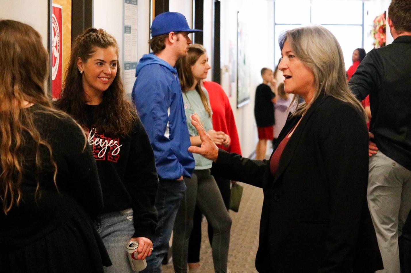 Photograph of a group of people engaged in conversation in a hallway, likely at a social or networking event. The scene features a woman with gray hair speaking animatedly to a younger woman holding a can, with others casually interacting in the background.