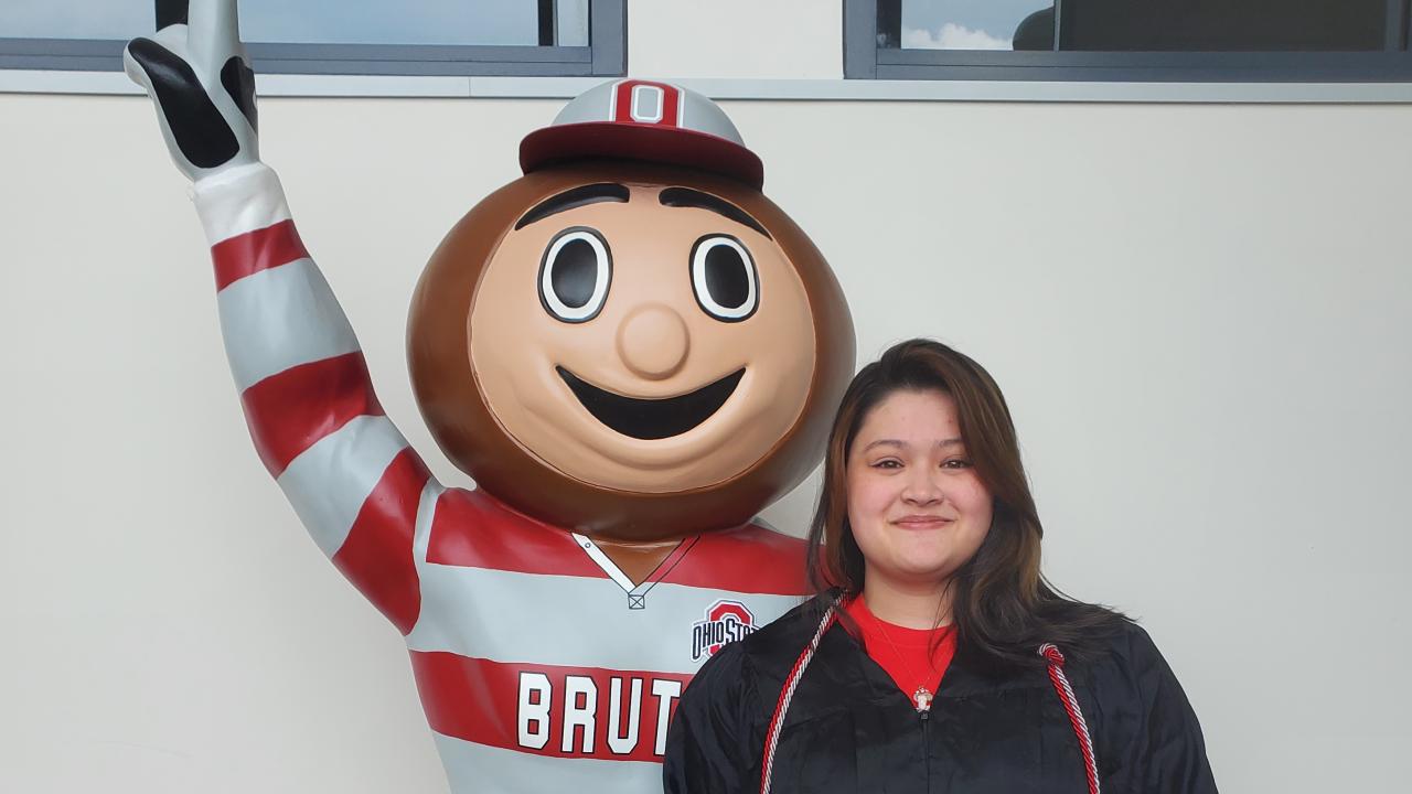 Female smiling displaying a diploma while standing next to Brutus Buckeye.